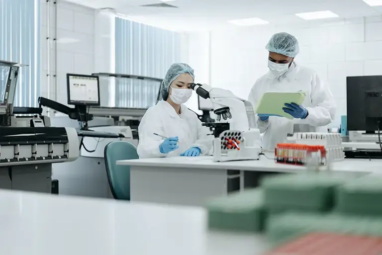 Two scientist are working in lab. One is seated and making notes after looking down her microscope, while her colleague stands nearby and he makes notes on a clipboard.